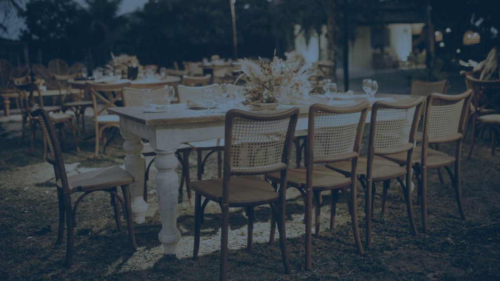Outdoor event dining setup with rustic tables, wooden cane-back chairs, and floral centerpieces, presented with a cool blue color filter.