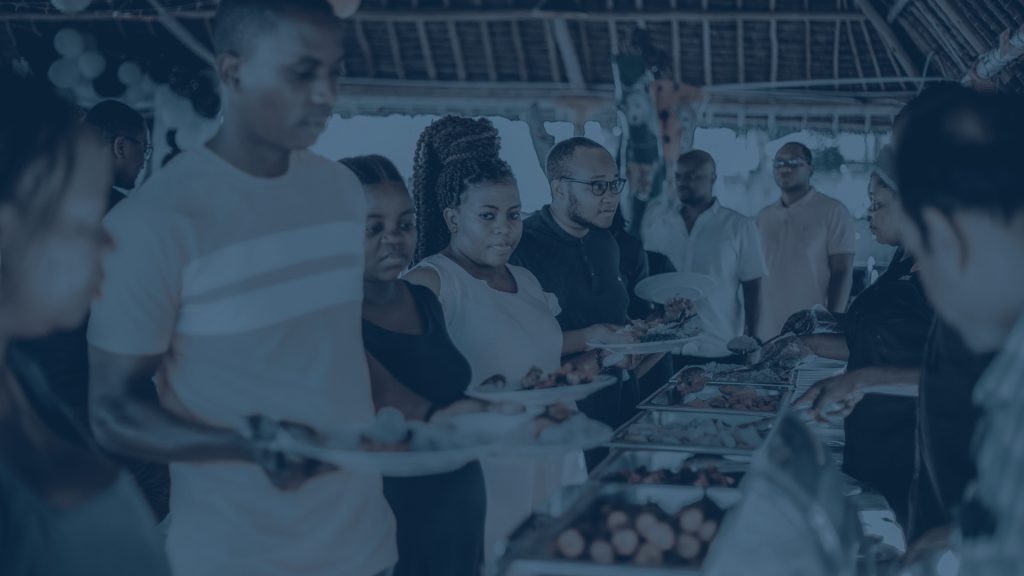 Event guests holding plates and serving themselves food from chafing dishes at a buffet line, under a covered structure; image has a blue color overlay.