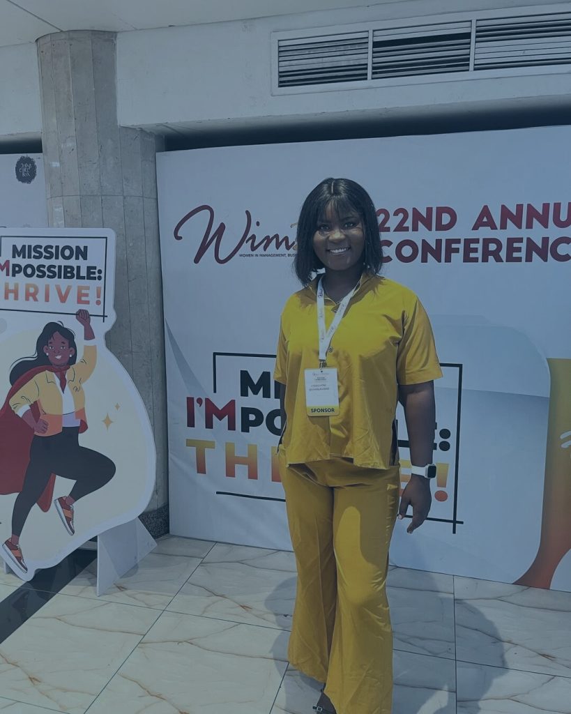 Woman in a yellow outfit smiling, wearing a 'SPONSOR' lanyard, poses at the Women in Management (WIMBIZ) 22nd Annual Conference in front of event backdrops.