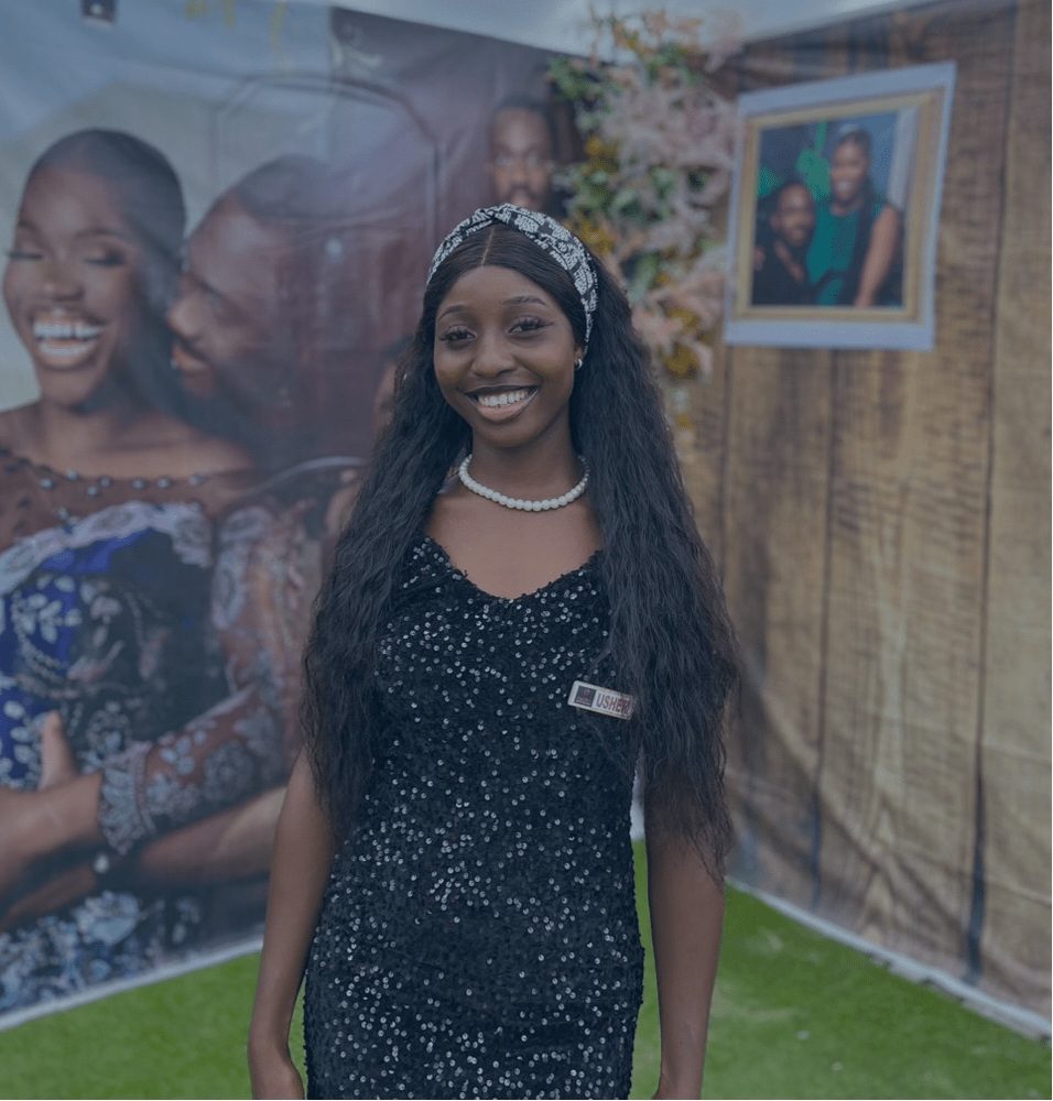 Female event usher smiling warmly, wearing a black sequined dress, pearl necklace, headband, and an 'USHER' badge, standing at an event.