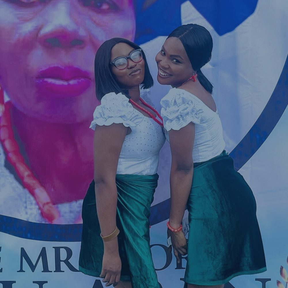 Two smiling ushers posing together in matching white tops, green skirts, and red beaded necklaces in front of an event backdrop.