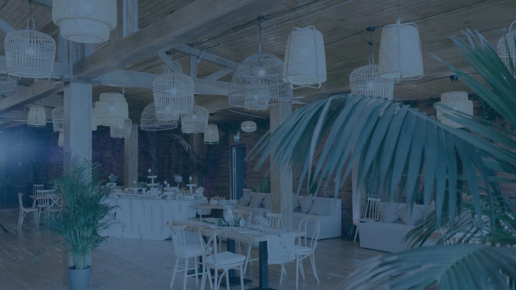 Interior of an event venue decorated with numerous woven hanging light fixtures, set dining tables, lounge seating, and plants, presented with a blue color overlay.