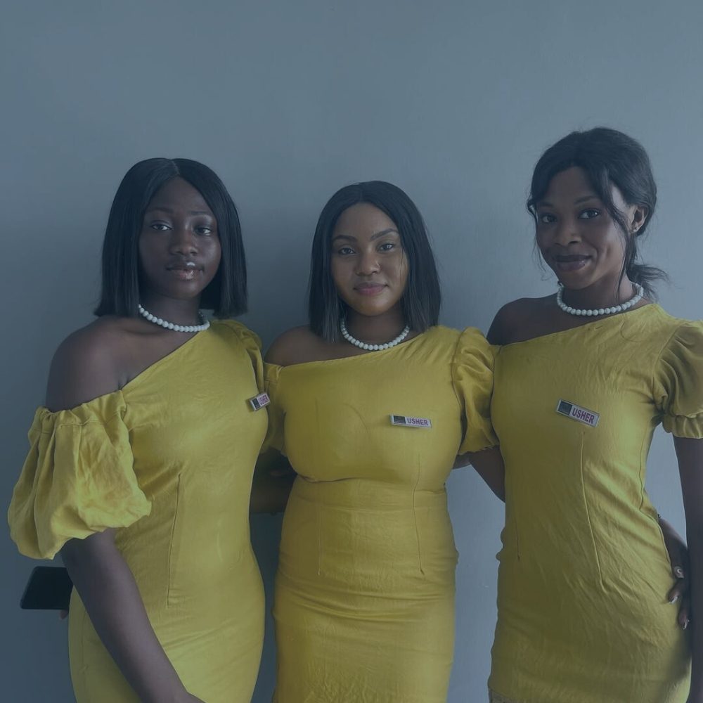 Three female event ushers posing together in matching yellow one-shoulder dresses, pearl necklaces, and 'USHER' badges against a grey background.
