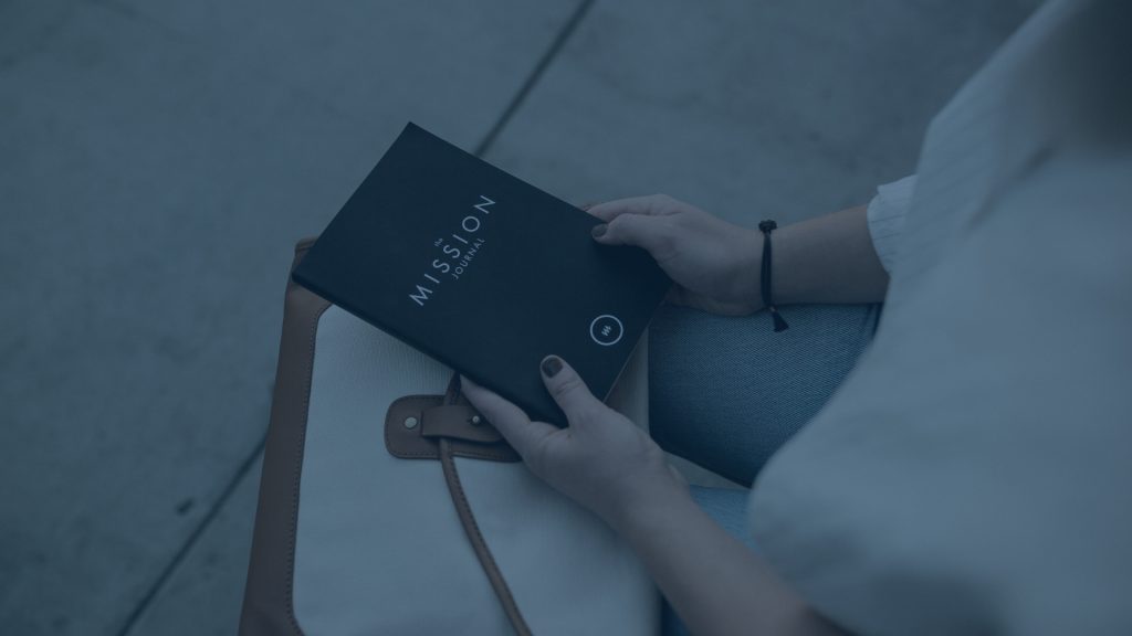 Person holding a black notebook titled 'the Mission Journal' while sitting, with a tote bag beside them; image has a cool blue tint.