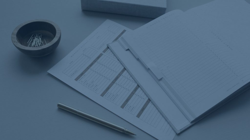 Budgeting tools laid out on a desk: 'Budget Overview' sheet, open notebook with 'Finance Tracker' pages, pen, and bowl of paperclips.