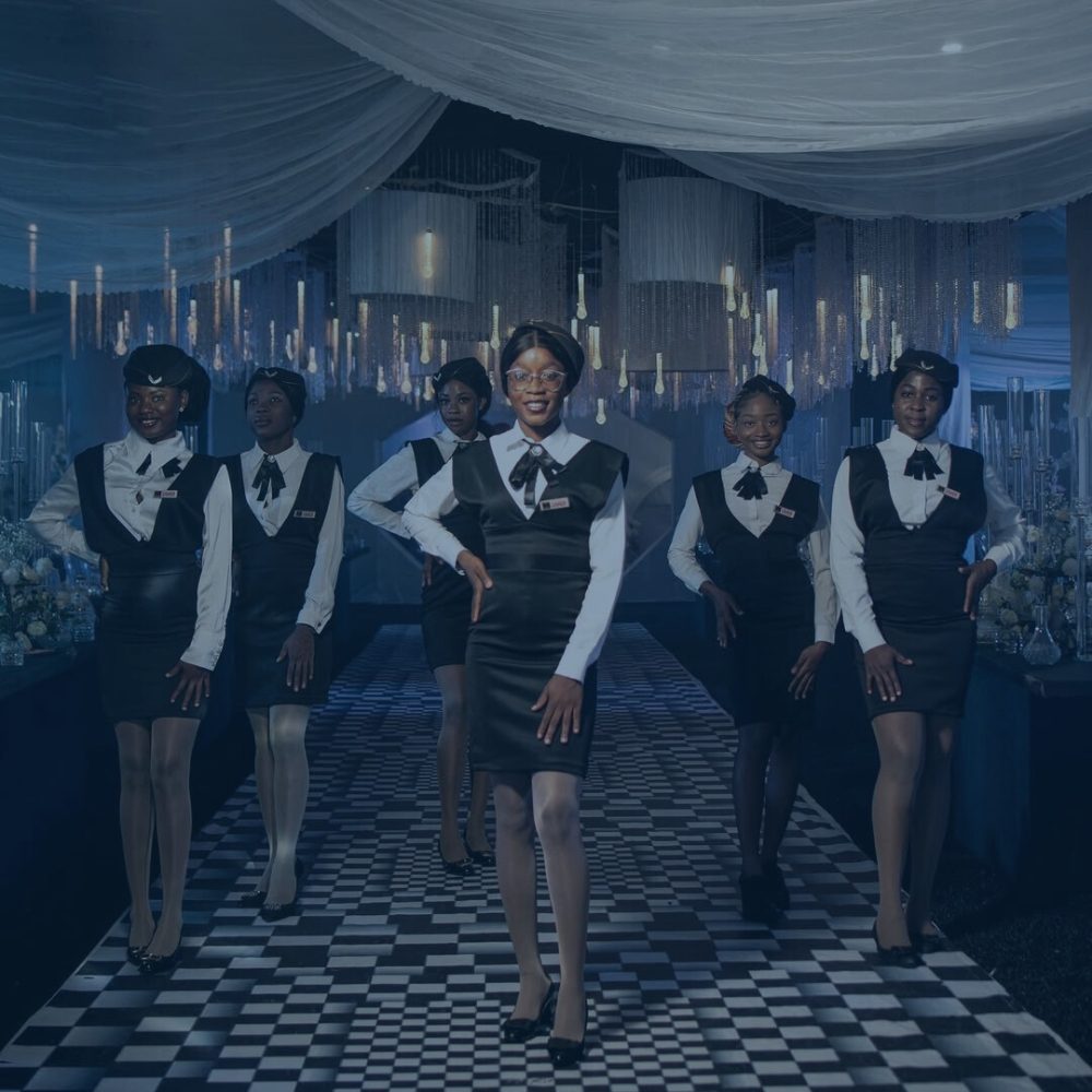 Six female event ushers in matching uniforms pose confidently on a black and white checkered floor in an elegantly decorated venue with hanging lights.